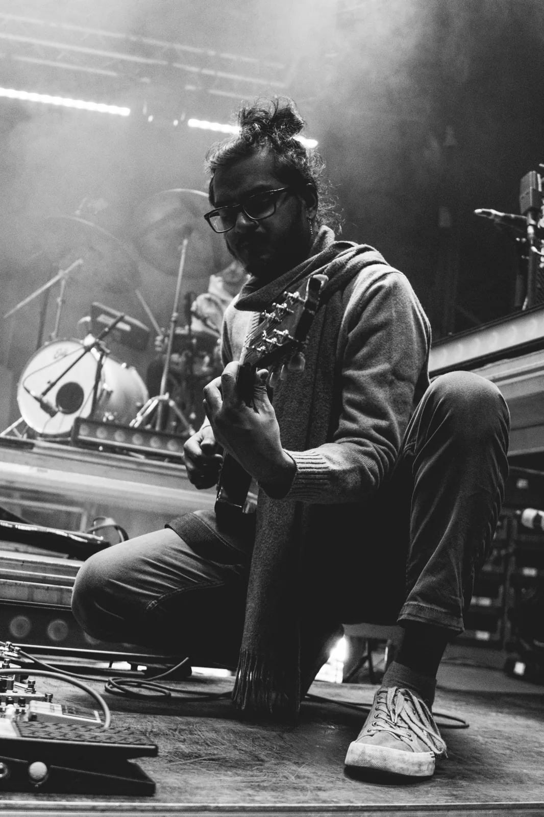 Black-and-white portrait of Ravi Ramsahye kneeling with his guitar, stage lights behind him