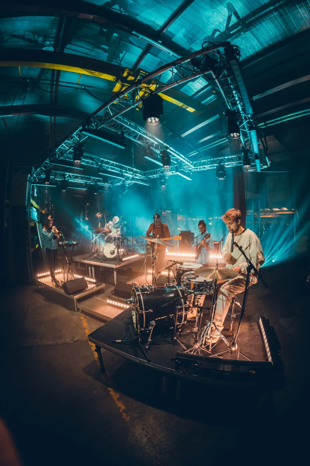 Ravi Ramsahye's band performing live on an industrial-style stage lit in blue and teal, with a trumpet player, drummer, guitarist, and bassist