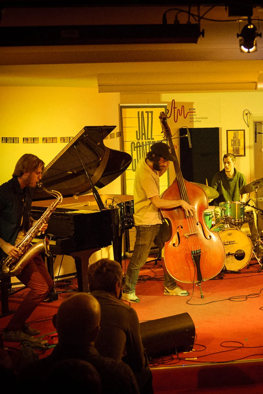 Wide shot of the full band performing live in a small club — saxophone, piano, upright bass and drums on stage under warm stage lights