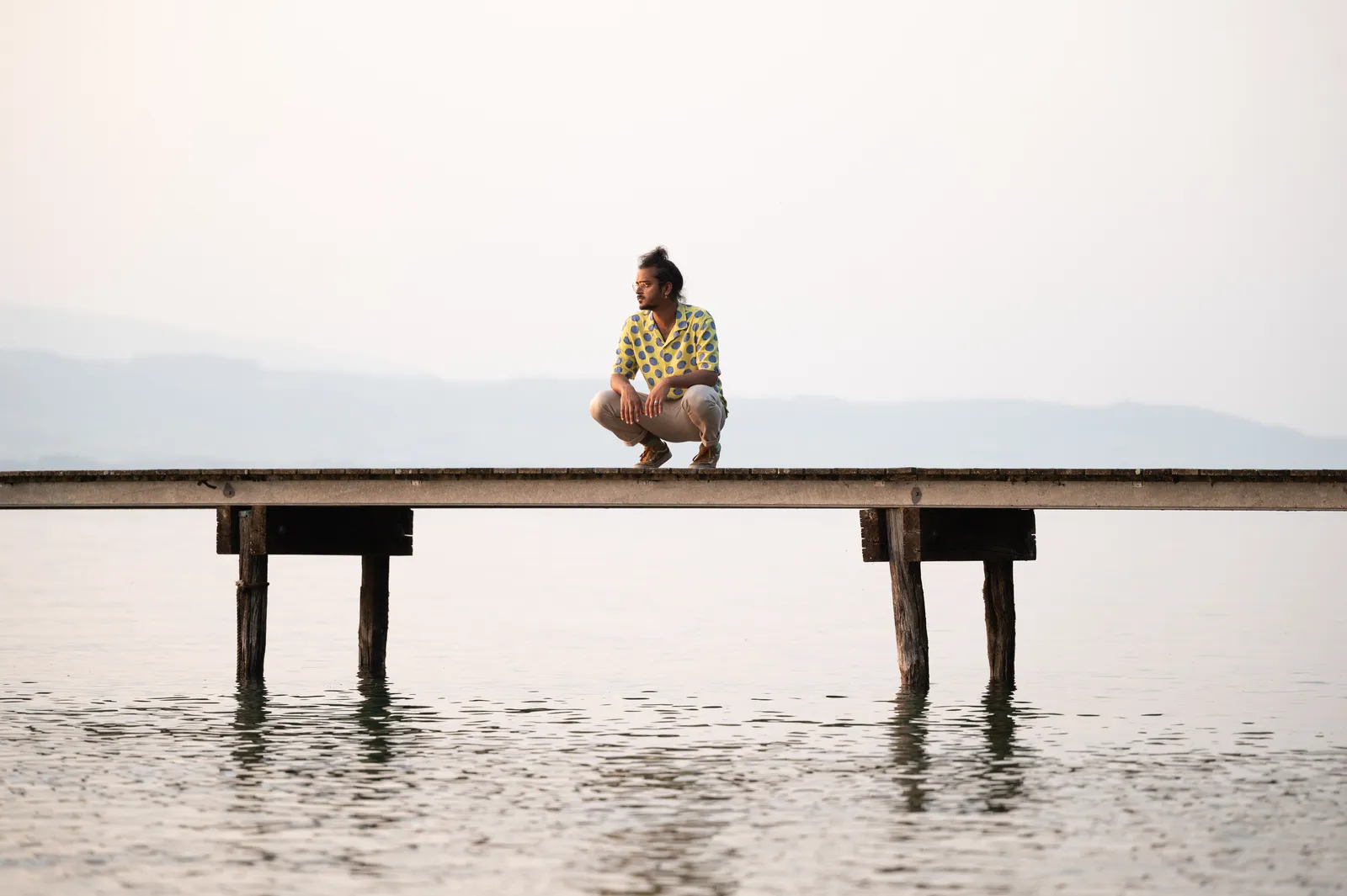 A lone figure sitting at the far end of a long pier over still water at dusk