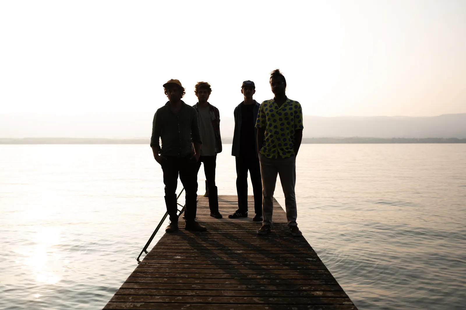 The four musicians of PROTOTYPE standing in silhouette at the end of a wooden jetty against a bright hazy lake at sunset